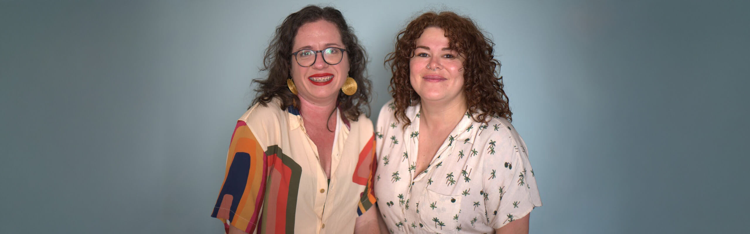 Retrato de duas mulheres sorrindo diante de um fundo azul-claro. À esquerda está Jade Azevedo, com cabelos escuros ondulados, óculos redondos, brincos grandes dourados e camisa clara com estampas geométricas coloridas. À direita está Janaina Moraes, com cabelos ruivos cacheados, piercing no nariz e camisa clara com estampas pequenas de palmeiras. Ambas estão posicionadas lado a lado, olhando para a câmera.