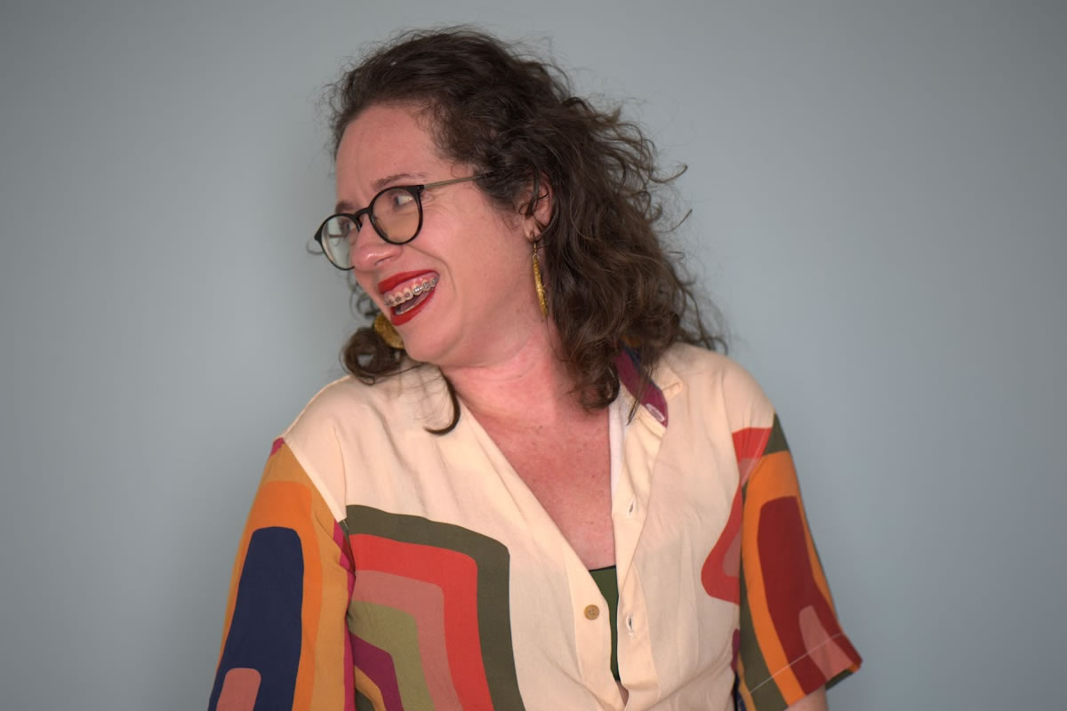 Retrato de Jade Azevedo sorrindo e olhando para a esquerda, em frente a um fundo liso azul-claro. Ela tem cabelos cacheados castanhos, usa óculos de armação escura, brincos longos e aparelho ortodôntico. Veste uma camisa clara com estampas geométricas coloridas em tons de laranja, vermelho, verde e azul. A fotografia transmite espontaneidade, alegria e descontração.